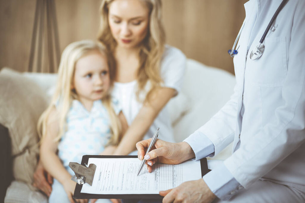 Doctor and patient. Pediatrician using clipboard while examining little girl with her mother at home. Sick and unhappy child at medical exam. Medicine concept - Photo, image
