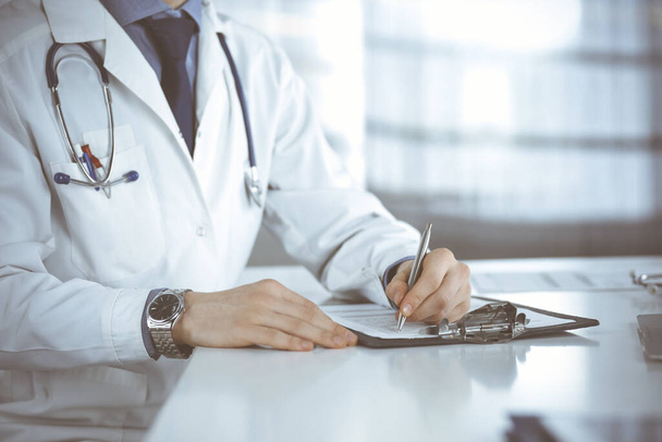 Unknown male doctor sitting and working with clipboard of medication history record in clinic at his working place, close-up. Young physician at work. Perfect medical service, medicine concept - Photo, Image
