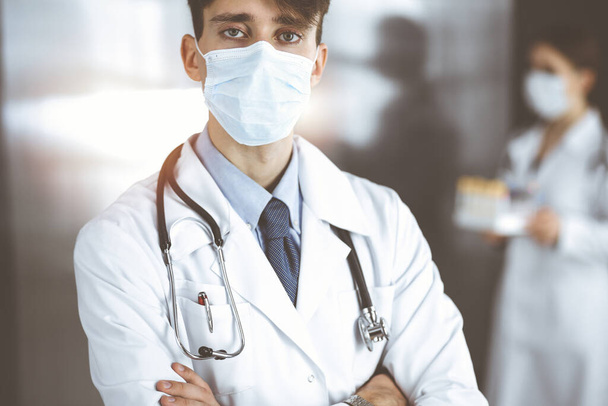 Young male doctor standing with arms crossed in clinic. Smart physician wearing medical protective mask in modern hospital. Medicine and Corona virus concept - Fotoğraf, Görsel
