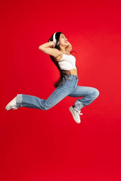 Asian woman with headphones jumping over red studio background with copyspace - Фото, зображення
