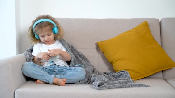 Little blonde girl, at home with headphones and with a mobile phone listens to music on the couch - Footage, Video
