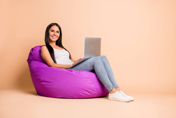 Full size profile photo of optimistic brunette girl sit pouf type laptop wear white top jeans sneakers isolated on beige background - Foto, afbeelding