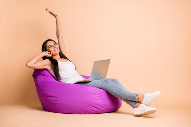 Full size profile photo of optimistic brunette girl sit pouf hold laptop wear white top jeans sneakers isolated on beige background - 写真・画像