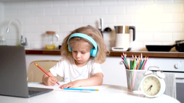 Niña rubia en los auriculares y delante de un ordenador portátil en casa en la cocina dibuja, el concepto de aprendizaje en línea - Metraje, vídeo