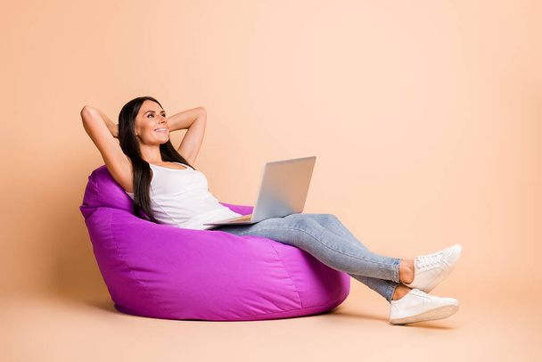 Photo portrait full body view of dreamy woman with laptop sitting in beanbag chair isolated on pastel beige colored background - Фото, зображення