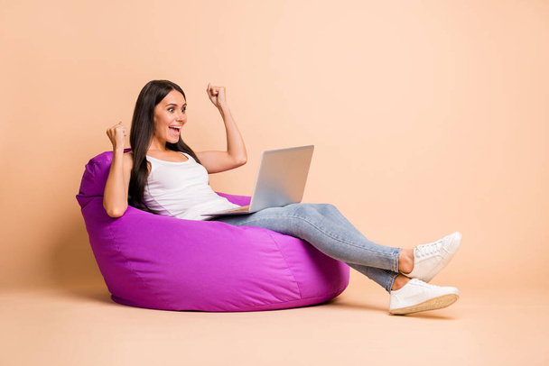 Full size profile photo of optimistic brunette girl sit pouf hands fists hold laptop wear top jeans isolated on beige background - Φωτογραφία, εικόνα
