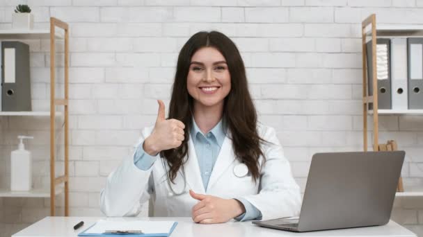 Mujer doctor gestos pulgares hacia arriba posando en el lugar de trabajo interior - Imágenes, Vídeo