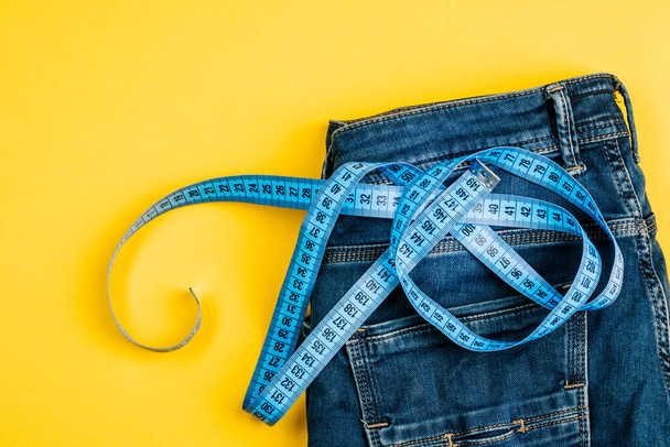 The concept of a healthy lifestyle and diet. Blue jeans with a blue measuring tape instead of a belt. - Фото, зображення