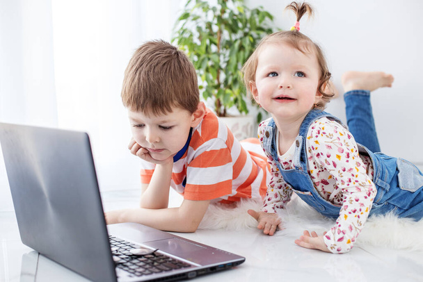 Młodszy brat i siostra oglądają śmieszne filmy na laptopie. Połóż się na podłodze. Koncepcja rodziny, dzieciństwa, internetu i gier. - Zdjęcie, obraz