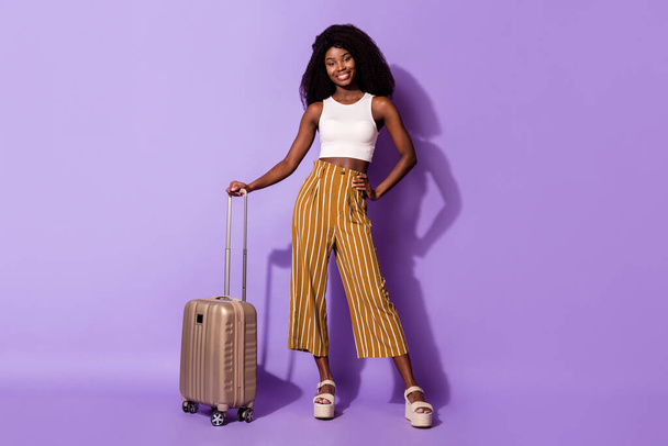 Full body photo of pretty cheerful dark skin lady put hand on waist hold suitcase isolated on violet color background - Zdjęcie, obraz