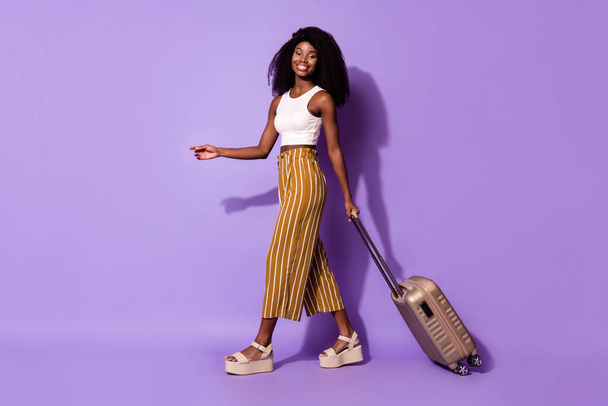 Full size profile photo of pretty positive dark skin girl arm hold suitcase look camera isolated on violet color background - Foto, Imagem
