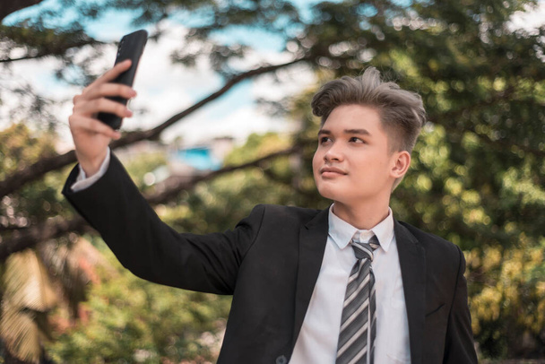 Un joven asiático seguro de sí mismo en traje de negocios y teñido de ceniza pelo rubio se toma una selfie de sí mismo mientras está en el parque. - Foto, Imagen