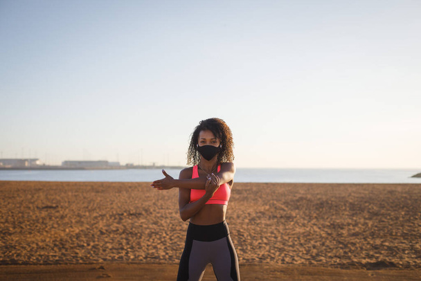 Young sporty black woman on fitness and running workout wearing face mask for protecting against coronavirus at city beach. Healthy lifestyle and exercising. Fit athlete stretching arms and shoulders for warming up.  Gijon, Asturias, Spain. - Fotografie, Obrázek