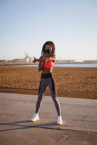 Young sporty black woman on fitness and running workout wearing face mask for protecting against coronavirus at city beach. Healthy lifestyle and exercising. Fit athlete stretching arms and shoulders for warming up.  Gijon, Asturias, Spain. - Foto, immagini