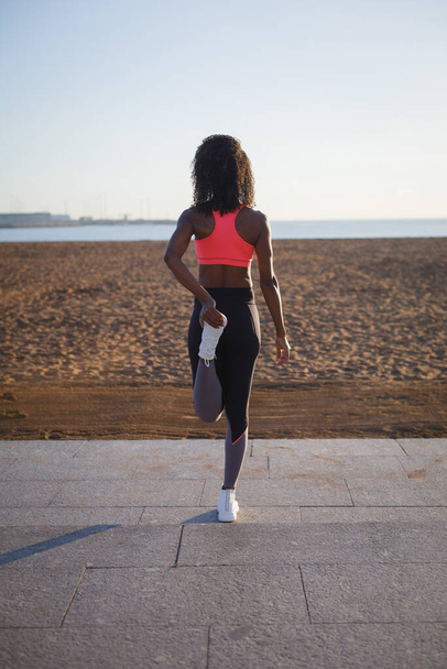 Young sporty black woman on fitness and running workout at city beach. Healthy lifestyle and exercising. Fit athlete stretching for warming up.  Gijon, Asturias, Spain. - Fotografie, Obrázek
