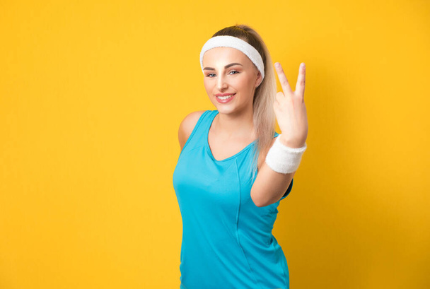Happy fitness young woman showing peace sign, isolated on yellow background. Half length of sportswoman ready for exercising. Athlete female person. 80s styling. Studio shot - Foto, Imagem