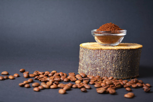 Πλευρική άποψη του καβουρδισμένου αλεσμένου καφέ σερβίρεται σε γυάλινο μπολ σε ξύλινο κορμό, με διάσπαρτους κόκκους καφέ σε σκούρο φόντο. Υψηλής ποιότητας σπόροι οργανικού αρώματος. Αντιγραφή εικόνας κενού κειμένου - Φωτογραφία, εικόνα