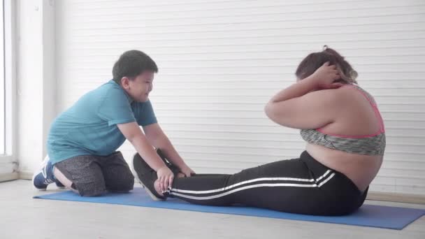 Famille asiatique avec la graisse de mère dans l'entraînement de vêtements de sport avec des sit-ups et l'aide de fils à la maison, l'exercice pour la perte de poids de régime, maman et garçon est l'activité d'obésité pour les soins de santé ensemble, à l'intérieur, plus la taille. - Séquence, vidéo