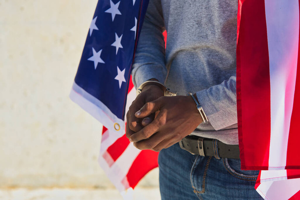 Αφροαμερικάνος με χειροπέδες στα χέρια του και σημαία των ΗΠΑ πάνω από τους ώμους του. Φυλετική καταστολή. - Φωτογραφία, εικόνα
