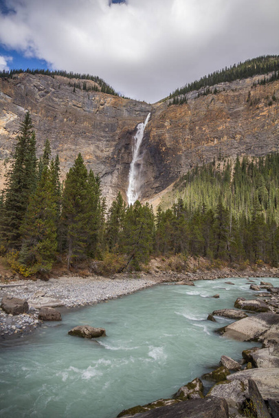 Sunlit water tumbles down Takakkaw Falls in Yoho National Park, BC.  A fast-flowing stream - turquoise with glacial silt - is in the foreground - Foto, imagen