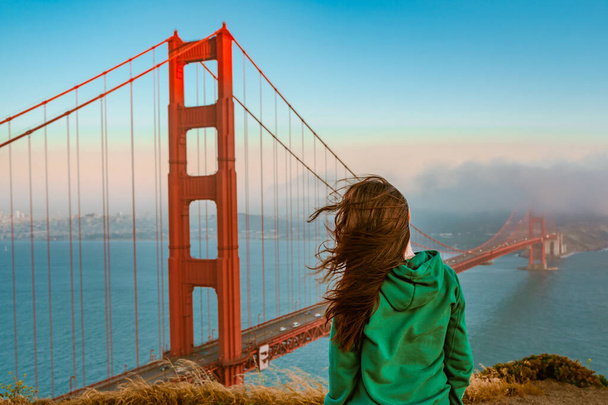 Eine junge Frau im grünen Kapuzenpulli steht bei Sonnenuntergang auf einem Hügel über der Golden Gate Bridge in San Francisco - Foto, Bild