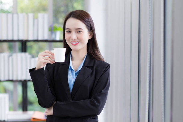 asiatico affari donna che indossa blu camicia e nero abito tiene caffè tazza in il suo mano e sorriso felicemente a lavoro ufficio come sfondo. - Foto, immagini