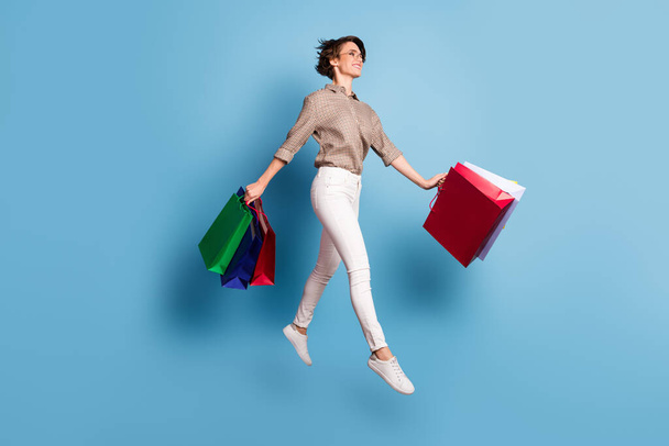 Full size profile portrait of cheerful pretty lady jumping hands hold bags isolated on blue color background - Фото, изображение
