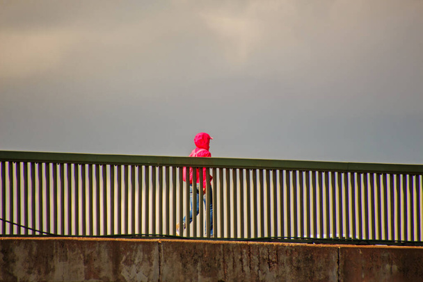a man in a pink jacket walks across a bridge under a rain-covered sky - Φωτογραφία, εικόνα