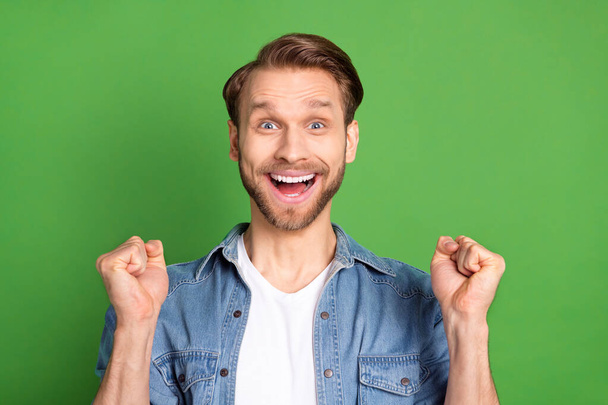 Photo portrait of young guy smiling in denim shirt gesturing like winner isolated on bright green color background - Фото, зображення