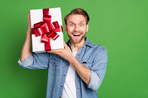 Photo portrait of young guy smiling in casual outfit trying to guess what in gift box isolated on vivid green color background - Foto, imagen