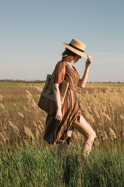 Çizgili elbiseli ve çayırda duran hasır şapkalı güzel bir kadın. Gündelik yaz modası, doğal ışıklandırmalı açık hava portresi.. - Fotoğraf, Görsel