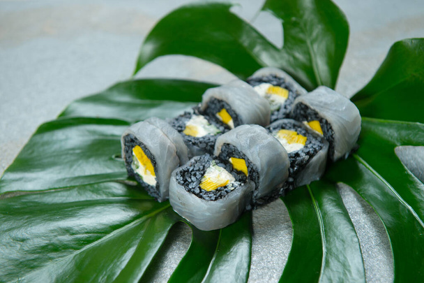 Κοντινό πλάνο στο ιαπωνικό μαύρο ρύζι ρολό σούσι με μελάνια ψαριού βοοειδών. Αγγούρι, ομελέτα, τυρί κρέμα τυλιγμένο σε ρύζι με φέτες κρέατος πετροψαριού από πάνω. Ασιατικό πιάτο σερβιρισμένο σε φύλλα τροπικών φυτών - Φωτογραφία, εικόνα