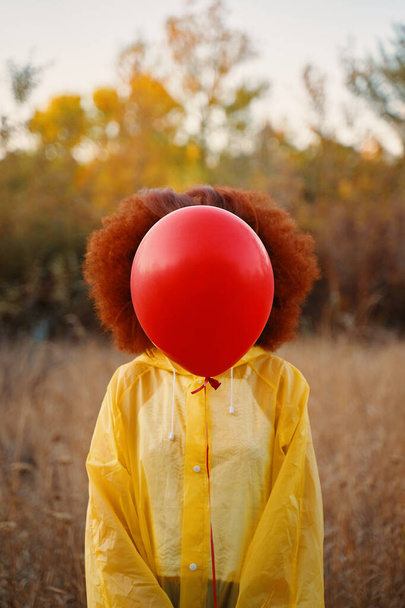 Sarı yağmurluklu bir kadın ormanın arka planında kırmızı bir balon tutuyor.. - Fotoğraf, Görsel
