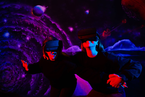 Две подружки в виртуальных очках держатся за руки в игровой комнате в неоновом свете.  - Фото, изображение
