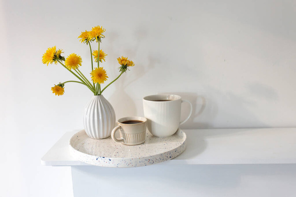 A bouquet of dandelions in a white fluted vase and two cups of different sizes with coffee on a white table. Breakfast. Copy space - Φωτογραφία, εικόνα