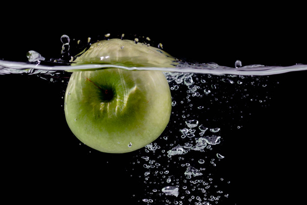 Reifer grüner Apfel fällt mit Spritzern vor schwarzem Hintergrund ins klare Wasser - Foto, Bild