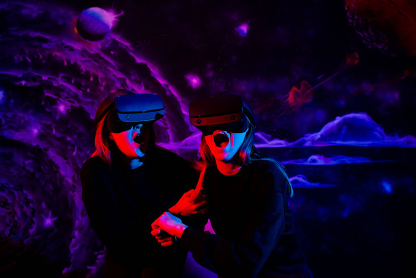 Due ragazze amiche sorelle con occhiali virtuali che si tengono per mano si divertono nella sala giochi a luce blu e rossa al neon.  - Foto, immagini