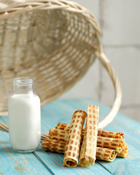 Wafer rolls or tubes and milk in glass jar on wooden table. Sweet rolls filled or stuffed with condensed milk. Soft focus. Vertical format - Φωτογραφία, εικόνα