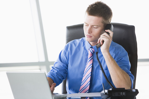 Businessman sitting in office with laptop on telephone - Foto, imagen