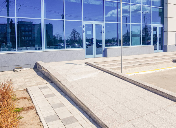 Close-up da rampa na entrada de um edifício moderno com uma fachada de vidro azul e concreto, reflexo do parque de estacionamento da cidade, acessibilidade para deficientes. - Foto, Imagem