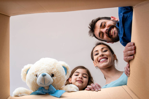 дом, люди, дети, переезд и концепция недвижимости - счастливая улыбающаяся семья открывает картонную коробку и распаковывает вещи в новом доме. - Фото, изображение
