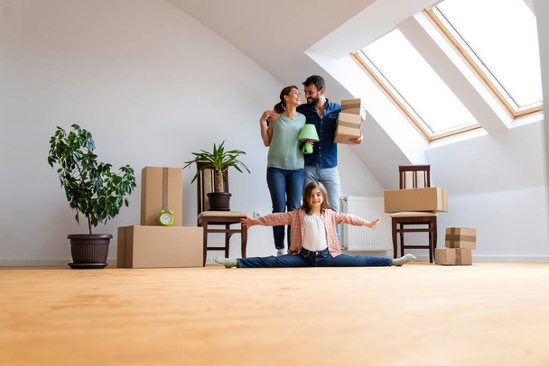 Porträt einer glücklichen kaukasischen Familie, die in eine neue Wohnung zieht, mit einem flexiblen kleinen Mädchen, das Gymnastik macht und Eltern, die Habseligkeiten tragen. - Foto, Bild