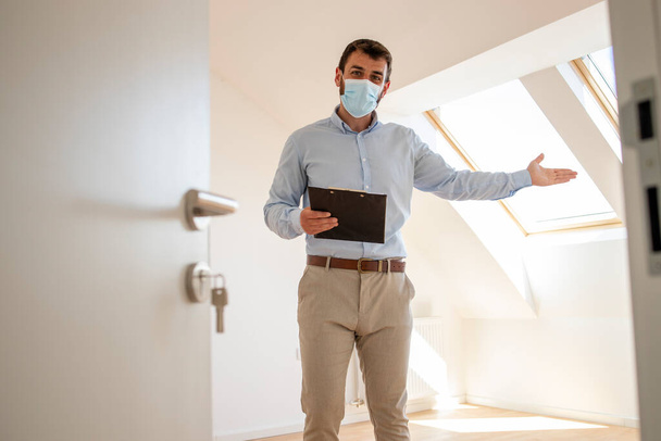 Porträt eines männlichen Immobilienmaklers mit Gesichtsmaske, der während der Coronavirus-Pandemie in einem neuen Haus steht. - Foto, Bild