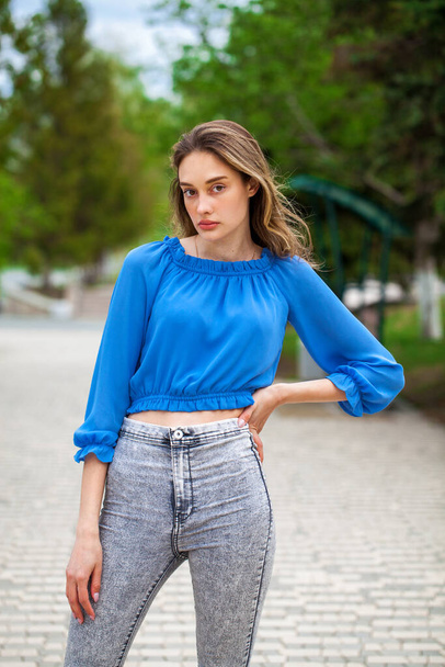 Young beautiful brunette girl in a blue blouse and gray jeans, summer park outdoor  - Foto, afbeelding