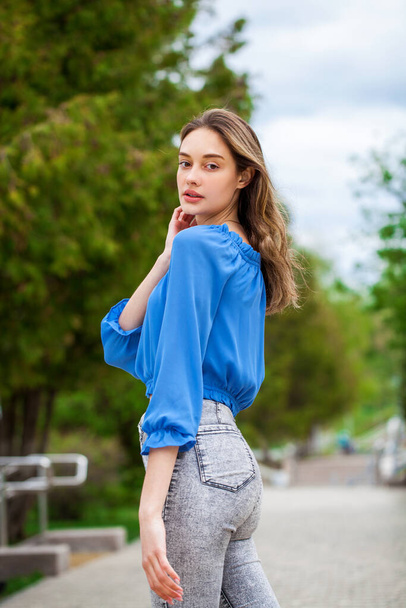 Young beautiful brunette girl in a blue blouse and gray jeans, summer park outdoor  - Фото, зображення