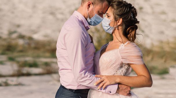 Giovane coppia amorevole a piedi in maschere mediche nel parco durante la quarantena il giorno del loro matrimonio. - Foto, immagini