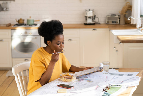 Занятый африканский рабочий или студент обедает в контейнере для еды на вынос, читает документ дома после работы - Фото, изображение