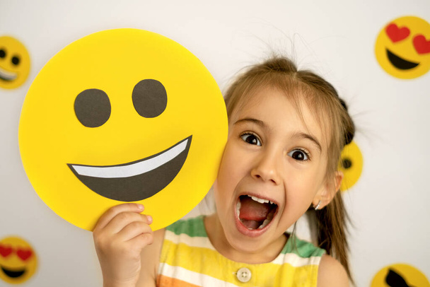 Wereldglimlachdag. Antropomorfe Smiley Gezicht. Een klein meisje met een glimlachend kartonnen lachend gezicht lacht hardop met haar mond open en haar ogen wijd en uitpuilend van verbazing. emoji dag - Foto, afbeelding