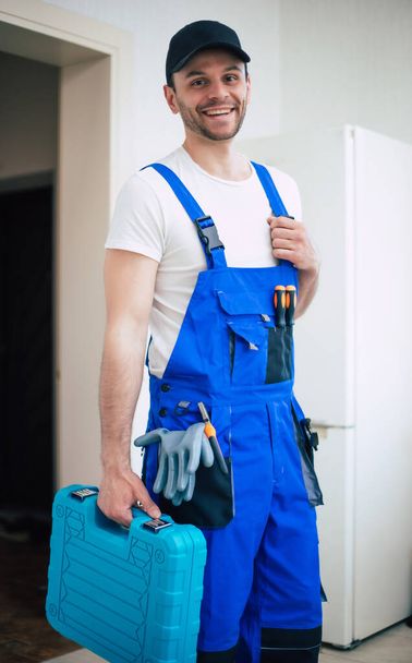 Professional young repairman in worker uniform and cap with modern toolbox with equipment after repair of refrigerator on the kitchen - Photo, image