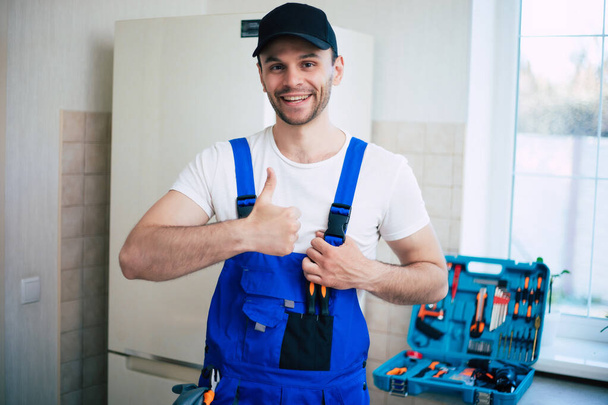 Professional young repairman in worker uniform and cap with modern toolbox with equipment after repair of refrigerator on the kitchen - Photo, image